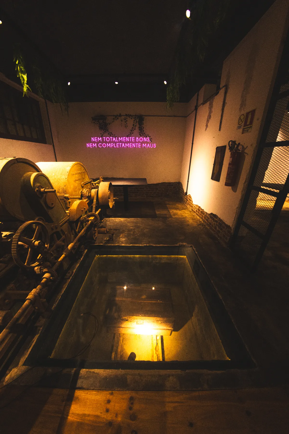 Interior da Cápsula: o maquinário têxtil original, o poço com piso de vidro iluminado por baixo e o letreiro de neon da casa ao fundo