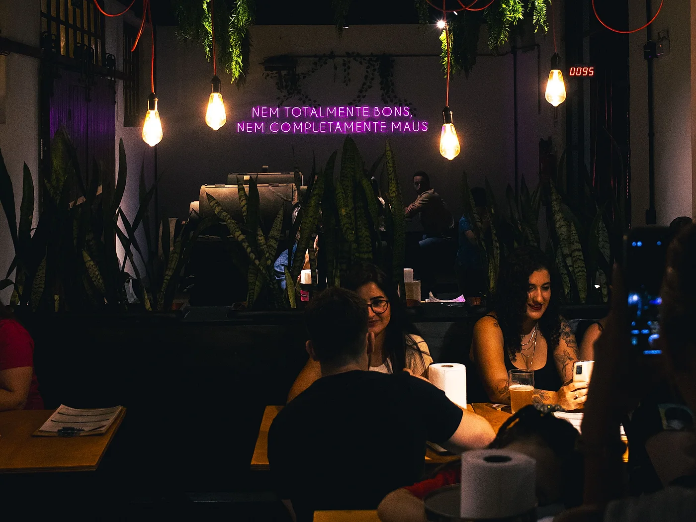 Salão da Cápsula à noite: lâmpadas penduradas, o letreiro de neon da casa e gente conversando nas mesas com chope