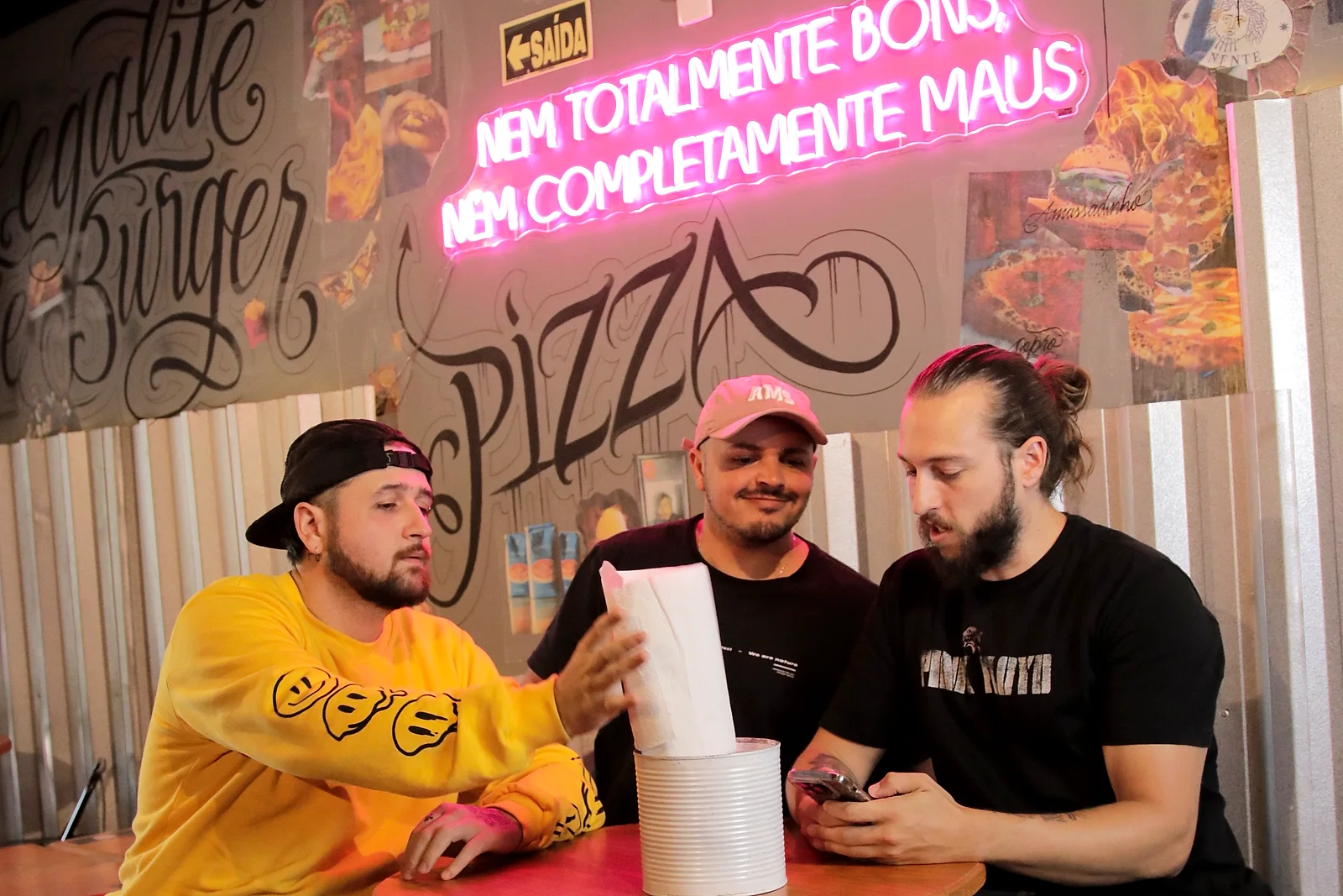 Os três sócios do PUGA à mesa, sob o letreiro de neon da casa: Caio à esquerda, Matheus no meio e Edu à direita